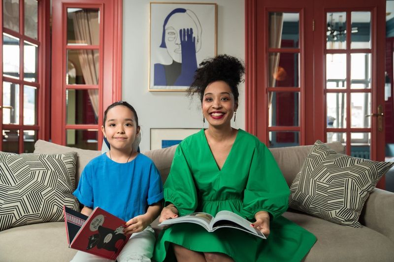 A mother in a green dress and her young daughter sit together on a sofa, each with an open book.