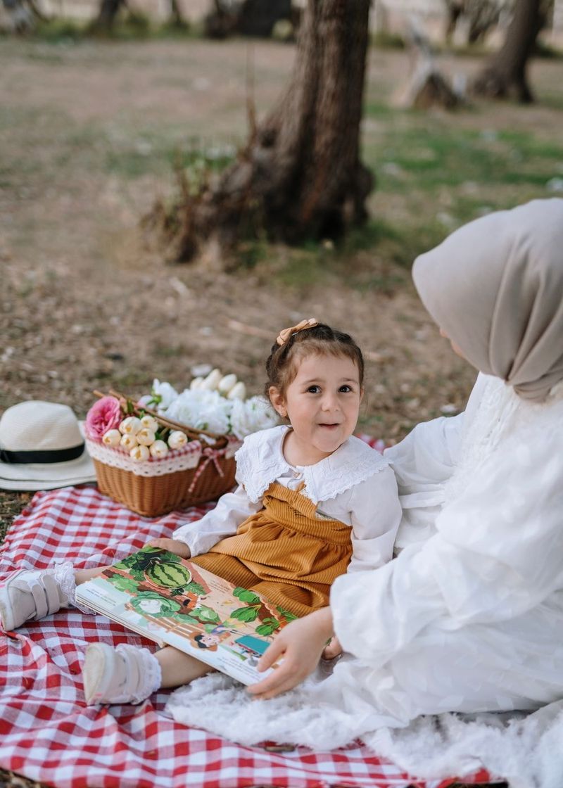 A small child with a picture book on a picnic blanket beside a parent in a headscarf.