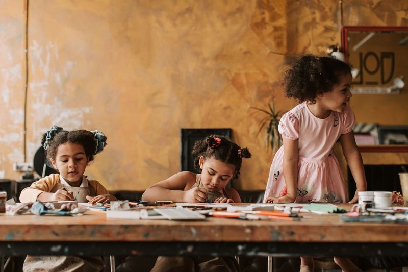 Children drawing and making at a long wooden table in a warm art studio.