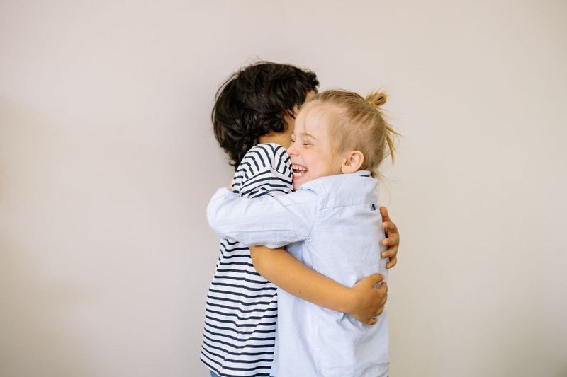 Two young children hugging each other tightly against a plain white wall.