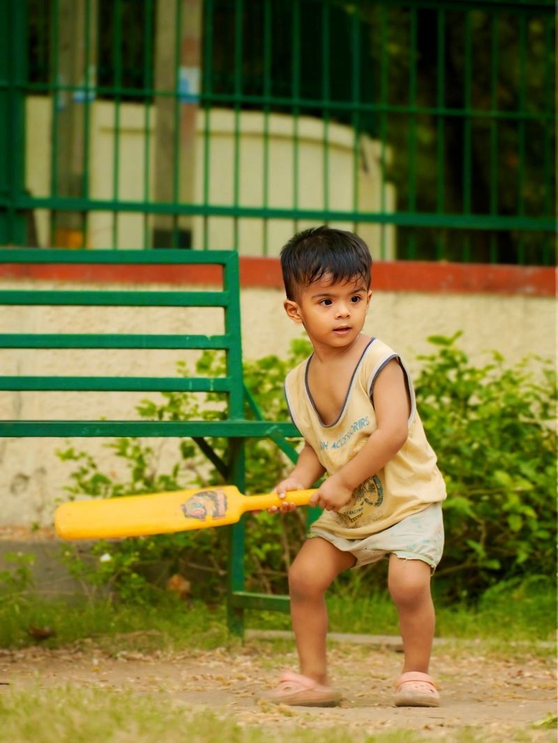 A toddler in a vest swinging a yellow plastic cricket bat in a park.