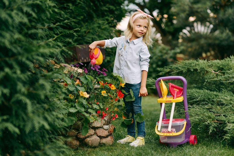 A little girl in a floral dress pouring water from a can in a flower garden, with a toy trolley beside her.