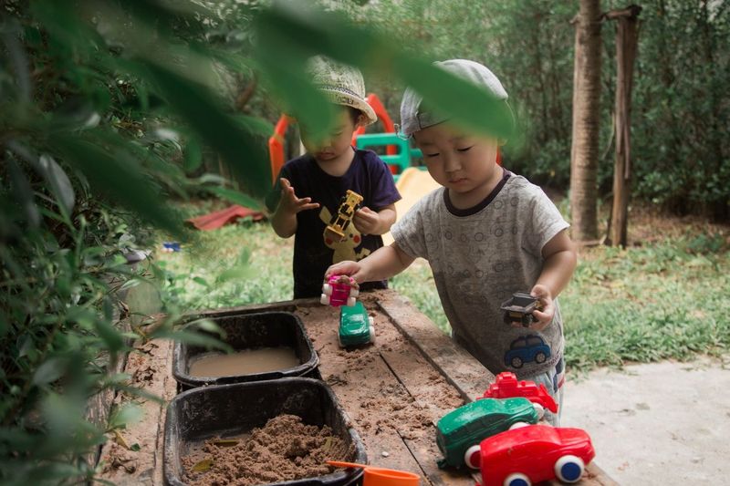 Two young boys playing at a wooden mud kitchen in a garden, scooping soil, with toy cars beside them.